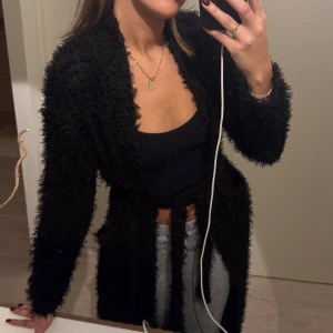 Svart fluffig set med kofta, topp och byxor - Säljer ett svart set med en fluffig kofta och matchande topp och byxor. Perfekt för en trendig och bekväm look. Koftan är lång och öppen framtill med knytband & fickor medan toppen är croppad och tight. Koftan är XS och byxor/tröja S, men allt passar en XS/S. Aldrig använd.