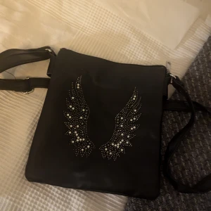 Svart axelväska med vingdetaljer - Snygg svart axelväska med coola vingdetaljer i form av små pärlor. Väskan har en justerbar axelrem och en praktisk dragkedja upptill. Perfekt för att ge din outfit en unik touch.
