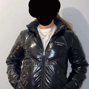 Svart pufferjacka från Calvin Klein Jeans - Snygg svart pufferjacka från Calvin Klein med glansig finish. Jackan har dragkedja framtill och en hög krage för extra värme. Perfekt för kyliga dagar. Slut på hemsidan i alla storlekar! Kontakta mig för fler bilder. Priset kan diskuteras vid snabb affär.🥰Nypris 2000!