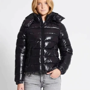 Svart pufferjacka - Snygg svart pufferjacka med dragkedja och hög krage. Perfekt för kyliga dagar med sin blanka yta och vadderade design. Jackan har långa ärmar och en figurnära passform.