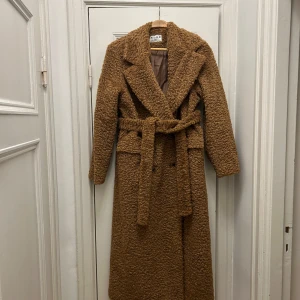 Brun teddyjacka från NA-KD - Säljer en mysig brun teddyjacka från NA-KD. Jackan har ett bälte i midjan och långa ärmar, perfekt för kyliga dagar. Den är lång och har en klassisk krage som ger en stilren look.