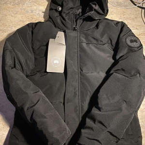 Canada Goose Jacka - Snygg svart dunjacka med huva och dragkedja. Perfekt för kalla dagar. Märkeslogga på ärmen ger en stilren touch.