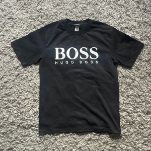 Hugo boss t shirt - Snygg svart t-shirt från Hugo Boss med stort vitt tryck på framsidan. Klassisk rund hals och korta ärmar. Perfekt för en stilren look.