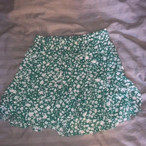 Grön blommig kjol - Säljer en superfin grön kjol med vita blommor. Det finns även inbyggda shorts!! Den är perfekt för sommaren med sitt lätta och luftiga material. Kjolen har en elastisk midja för en bekväm passform och en söt volangdetalj längst ner. Perfekt för en dag på stranden eller en somrig picknick! 🌸