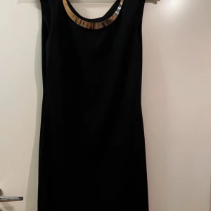 Svart klänning med guldhalsband - En enkel svart klänning med fina detaljer av guld på halsbandet som sitter fast på klänningen. Den är knälång och är i storlek 38.