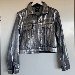 Silverfärgad jacka från Gina Tricot - Säljer en supercool silverfärgad jacka från Gina Tricot i samarbete med Icona Pop. Jackan har en glansig finish och klassisk jeansjacka-stil med knappar framtill. Perfekt för att sticka ut på festen eller bara för en extra touch av stil i vardagen. 🪩✨