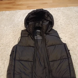 Svart dunväst från Zara - Säljer en svart dunväst från Zara med dragkedja och huva. Perfekt för kyligare väder med sin quiltade design. Västen är i storlek XS och har en stilren look.