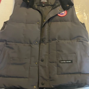 Svart dunväst från Canada Goose - Säljer en svart dunväst från Canada Goose med dragkedja och knappar. Västen har två stora fickor framtill och en broderad logotyp på bröstet. Perfekt för kyliga sommar nätter dagar.