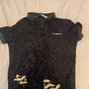 Svart pike från Off-White - Säljer en piké t-shirt från Off-White med ett unikt mönster och gula detaljer. T-shirten har korta ärmar och en klassisk krage. Perfekt för dig som vill ha något stilrent men ändå iögonfallande. Storlek m, pris kan diskuteras vid snabb affär. Made in Bangladesh. Qr kod finns