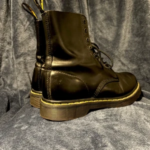 Dr. Martens  - Snygga svarta boots från Dr. Martens i skinn med klassisk gul söm runt sulan. Skorna har snörning och en robust design som passar perfekt för en cool och stilren look.