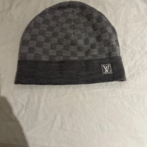 Grå rutig mössa från Louis Vuitton - Snygg grå mössa från Louis Vuitton med ett diskret rutigt mönster. Mössan har en broderad LV-logga på kanten och är perfekt för att hålla dig varm med stil. Tillverkad i ett mjukt och bekvämt material.