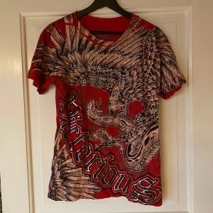 Röd t-shirt med tryck från Victorious - Snygg röd t-shirt från Victorious med ett stort, detaljerat tryck av en örn och texten 'Victorious' i svart och silver. Exakt lika kvalitet som när den köptes då den bara har använts ett par få gånger och inte längre passar mig