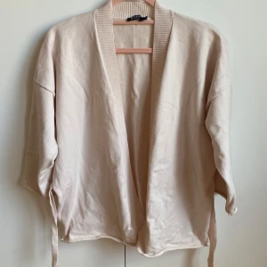 Beige kofta från Tiger of Sweden - Säljer en stilren beige kofta från Tiger of Sweden. Koftan har trekvartsärmar och ett knytband i midjan för en justerbar passform. Perfekt för en avslappnad och elegant look.