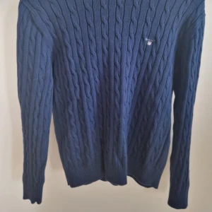Mörkblå stickad tröja från Gant - Säljer en stilren mörkblå stickad tröja från Gant med kabelstickat mönster. Tröjan har långa ärmar och en liten logotyp på bröstet. Perfekt för kyligare dagar. Passar både till jeans och chinos. Tröjan är i storlek 160