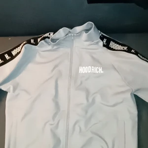 Grå hoodie från Hoodrich - Säljer en grå hoodie från Hoodrich med dragkedja och tryckt logga på bröstet. Tröjan har långa ärmar och svarta detaljer med text längs axlarna. Perfekt för en stilren look.
