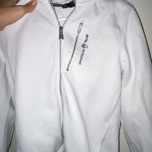 Vit jacka från Sail Racing - Snygg vit jacka från Sail Racing med dragkedja framtill och en bröstficka med dragkedja. Jackan har en stilren design med långärmade ärmar och en huva. Perfekt för en sportig look.