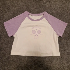 Vit och lila t-shirt med tennistryck - Säljer en söt vit och lila t-shirt med texten 'Tennis Ball Society' och två tennisrack på framsidan. T-shirten har korta ärmar och är ribbad i materialet. Perfekt för en avslappnad dag!