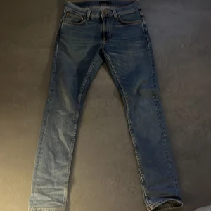 Blå jeans från Nudie Jeans - Snygga blå jeans från Nudie Jeans med klassisk femficksdesign och orange sömmar. Jeansen har en straight passform och är tillverkade i slitstarkt denim. Perfekta för en avslappnad stil.