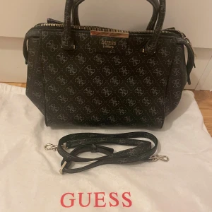 Svart handväska med mönster - Snygg svart handväska med mönster och dragkedja. Den är köpt för 1099 kroner och är ifrån Märket guess. de är lite suddigt bild men den är endast lite använd.