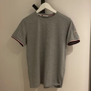 Grå t-shirt från Moncler - Snygg grå t-shirt från Moncler i slim fit. Den har korta ärmar med ribbade muddar i rött, vitt och blått. Perfekt för en stilren look.