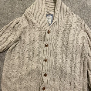 Beige stickad kofta  - Säljer en  beige stickad kofta. Koftan har ett klassiskt flätmönster och en bred krage som ger extra värme. Perfekt för kyliga dagar!