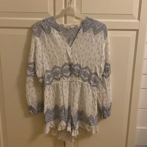 Vit och blå  playsuit från Ghanda - Charmig vit playsuit med blått mönster från Ghanda. Playsuiten har en v-ringad hals och långa ärmar. Den är dekorerad med ett geometriskt mönster och har volangdetaljer vid nederkanten. Perfekt för en bohemisk look! Storlek M. I fint skick. 