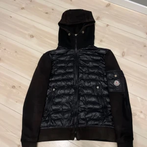Svart jacka från Moncler - Säljer en snygg svart jacka från Moncler med quiltad design och huva. Jackan har en dragkedja framtill och en ficka med knappdetalj på ärmen. Perfekt för kyligare dagar.mer bilder kom privat.