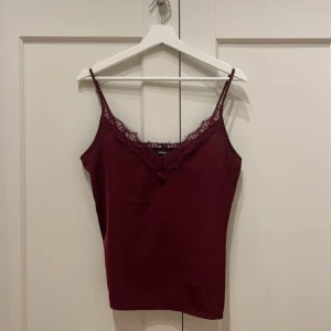 Vinröd Only  linne med spetsdetaljer - Säljer ett stilrent vinrött linne med smala axelband och vacker spetsdetalj vid halsringningen. Perfekt för lager-på-lager eller som det är. Passar till både vardag och festliga tillfällen.