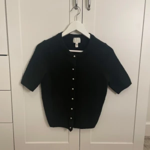 Svart kofta från H&M - Snygg svart kofta från H&M med korta ärmar och dekorativa pärlknappar framtill. Använd fåtal gånger så den är i väldigt bra skick. Jag skickar även med en extra knapp som jag fick med när jag kopte den om någon av knapparna skulle trilla av.