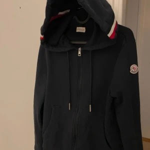 Mörkblå hoodie med dragkedja från Moncler - Säljer en snygg mörkblå hoodie från Moncler med dragkedja och huva. Den har en broderad logga på ärmen och ränder i rött och vitt på huvan. Perfekt för en stilren och bekväm look. Den är i storlek M.