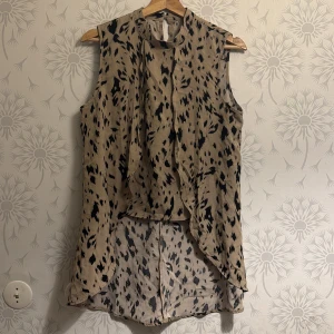 Beige ärmlös klänning med mönster - Snygg beige ärmlös klänning med svart leopardmönster. Den har en asymmetrisk design med längre bakstycke och en stilren krage. Perfekt för en trendig look.
