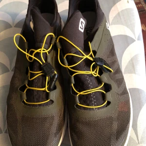 Svarta och gröna sneakers från Salomon - Snygga svarta och gröna sneakers från Salomon med gula snören och en robust sula. Perfekta för utomhusaktiviteter med en modern design och bekväm passform.