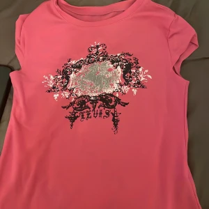 Rosa t-shirt med tryck - Säljer en rosa t-shirt med ett dekorativt tryck i svart och silver på framsidan. T-shirten har korta ärmar och en rund halsringning. Perfekt för en avslappnad stil.