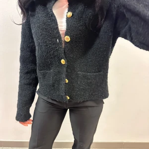 Svart stickad kofta med guldknappar - Säljer en snygg svart stickad kofta med iögonfallande guldknappar. Koftan har en normal passform och långa ärmar, perfekt för kyligare dagar. Den är både stilren och bekväm, ett måste i garderoben!