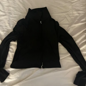 Svart ribbad tröja med dragkedja - Snygg svart tröja med ribbad struktur och dragkedja framtill. Tröjan har en hög krage och långa ärmar, perfekt för en stilren look. Passar bra till både vardag och mer uppklädda tillfällen.