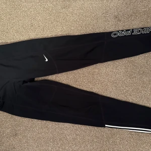 Svarta träningsleggings från Nike - Snygga svarta träningsleggings från Nike med vit logga och texten 'NIKE PRO' längs benet. Perfekta för träning med en tight passform och elastiskt material som ger bra rörelsefrihet. Fint skick med ett litet slitage som syns på sista bilden💛