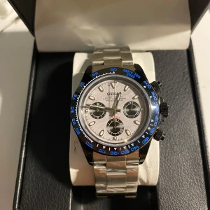Ny Seiko Daytona, ”Seikona” Meteorite Mod - Snygg Seiko herrklocka med silverfärgad länk och blå detaljer på boetten. Urtavlan är vit med tre mindre svarta subdials och tydliga markeringar. Perfekt för den som vill ha en stilren och sportig look. Urverket är quartz.