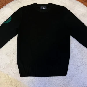 Svart tröja från Fred Perry - Säljer en stilren svart tröja från Fred Perry med ett grönt broderat märke på ärmen. Tröjan har långa ärmar och är tillverkad i ett mjukt material.