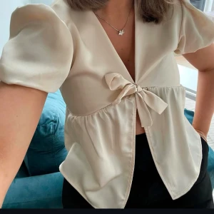 Beige blus med knytning - Säljer en söt beige blus med puffiga korta ärmar och knytning framtill. Perfekt för en stilren och elegant look. Blusen har en lätt v-ringning och en luftig passform.