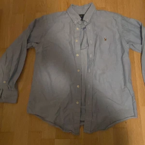 Oxford skjorta från Ralph Lauren - Säljer en klassisk blå skjorta från Ralph Lauren med broderad logga på bröstet. Skjortan har långa ärmar och knappar framtill. Perfekt för en stilren look. Ett litet hål på nacken men inte så märkvärdigt, funkar bra att sy igen 