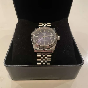 Stilrent Seiko-mod  - Just nu säljs en elegant Seiko-modifierad automatisk klocka med en mörkblå stilren urtavla som sticker ut från vardagen. Denna klocka har en unik design med noggrant utvalda detaljer som ger den en modern känsla. Såsom en räfflad bezel som ger en trendig look. Länkar ingår, vilket gör att du kan justera storleken för en perfekt passform. .