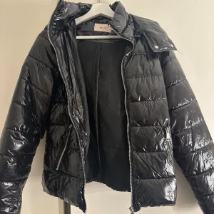 Svart glansig pufferjacka från Nelly - Snygg svart glansig pufferjacka från Nelly med dragkedja och huva. Jackan har en quiltad design och två praktiska fickor med dragkedjor. Perfekt för kyligare dagar när du vill hålla stilen. 🖤