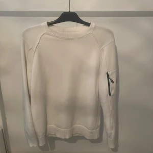 Vit tröja C.P. Company - Säljer en stilren vit tröja från C.P. Company. Tröjan har långa ärmar och en ribbad krage samt muddar. Detaljerad med en liten ficka på ärmen med dragkedja och en rund logotyp. Perfekt för en minimalistisk look.