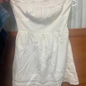 Vit studentklänning från Blanco - Charmig vit klänning från Blanco med vackra spetsdetaljer och smockad överdel. Perfekt för sommarens varma dagar. Klänningen har en luftig passform och är tillverkad i ett lätt material.