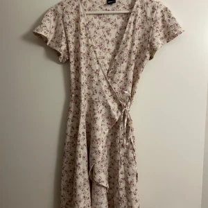 Blommig omlottklänning  - Fin sommrig omlottklänning från Gina Tricot!  Klänningen går till mitten av låren och har korta ärmar och ett band som man knyter i midjan🌸