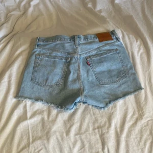 Ljusblå jeansshorts från Levi's - Säljer ett par ljusblå jeansshorts från Levi's med fransiga kanter och klassisk femficksdesign. Shortsen har en knappgylf och en läderpatch med logga baktill. Perfekta för en avslappnad stil.