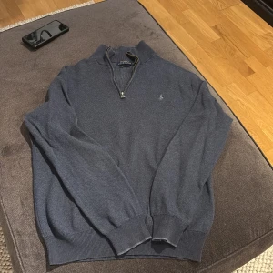 Blå tröja med dragkedja från Polo Ralph Lauren - Säljer en stilren blå tröja från Polo Ralph Lauren med dragkedja och broderad logga på bröstet. Tröjan har långa ärmar och ribbade muddar vid ärmslut och nederkant. Perfekt för en avslappnad men snygg look.