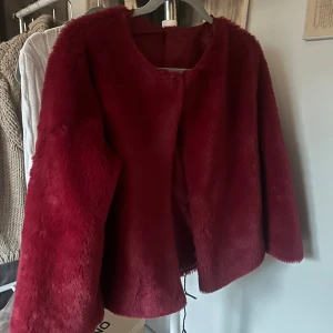 Vinröd pälsjacka - Säljer en snygg vinröd pälsjacka med långärmad design. Jackan har en mjuk och fluffig känsla, perfekt för att hålla dig varm och stilren under kyliga dagar. Den är öppen framtill utan knappar eller dragkedja.