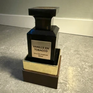 Vanille en Tobacco Eau de Parfum 80ml - Elegant parfym i mörkbrunt glas med en lyxig guldig etikett. Doften kombinerar vanilj och tobak för en sofistikerad och varm upplevelse. Flaskan har en unik fyrkantig design med en matchande förpackning i brunt och guld, 90-95% lik orginalet Tom ford Tobacco Vanille