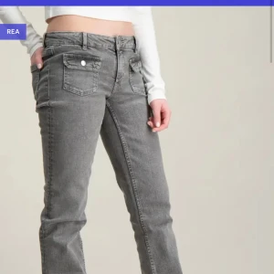 Grå jeans från gina young  - Snygga grå jeansbyxor med bootcut-stil och lågmidja. Byxorna har framfickor med lock och knappar, vilket ger en cool detalj. Perfekta för en avslappnad look. Storlek 170